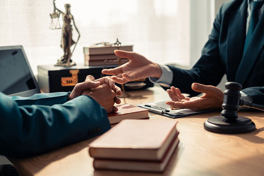 Two people in suits sit across a desk discussing legal matters. Their hands are visible, with books, a clipboard, a gavel, and a Lady Justice statue on the table—suggesting a Real Estate Attorney’s office in Macomb MI.