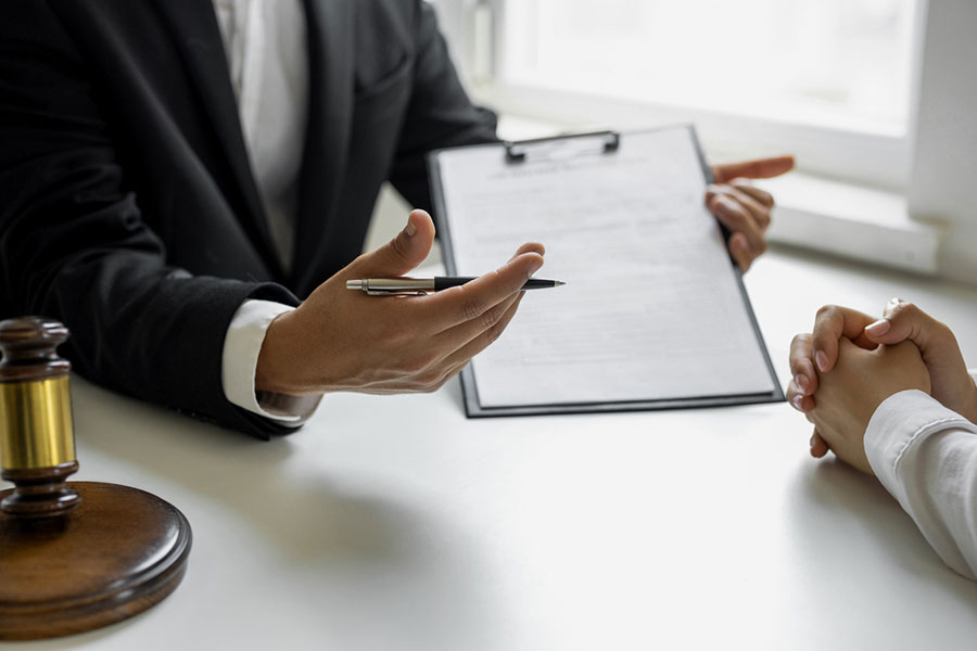 A Real Estate Attorney in a suit holds a clipboard and gestures with a pen while speaking to another person across a desk in Birmingham MI; a gavel on the desk suggests a legal or professional meeting.