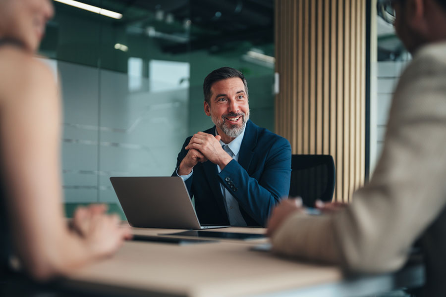 A man in a business suit sits at a desk with a laptop, smiling and talking to two blurred colleagues in a modern Oakland Charter Township office, reflecting the professionalism of an MI real estate attorney at work.