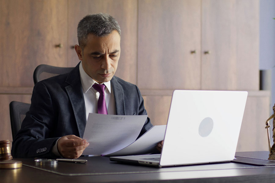 A Real Estate Attorney in Oakland Charter Township, MI, sits at a desk reviewing documents beside a laptop, with a gavel and Lady Justice statue suggesting a legal or office setting. A Real Estate Attorney in Oakland Charter Township, MI, sits at a desk reviewing documents beside a laptop, with a gavel and Lady Justice statue suggesting a legal or office setting.